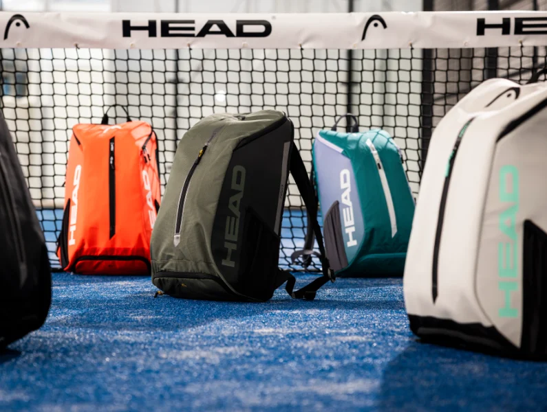 Vier HEAD Padel Rucksaecke in orange, khaki, tuerkis und weiss auf einem blauen Padelcourt vor dem Netz mit HEAD Banner - HEAD Padel Taschen Kollektion bei Padel4Players
