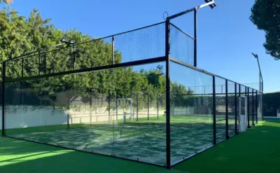Ein moderner Outdoor-Padel-Court mit Glaswänden, Metallrahmen und grünem Kunstrasen inmitten von Bäumen unter blauem Himmel.