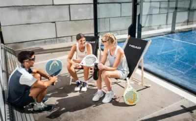 Drei Padel-Spieler sitzen entspannt am Spielfeldrand auf Liegestühlen, unterhalten sich und halten HEAD Padel-Schläger in den Händen.